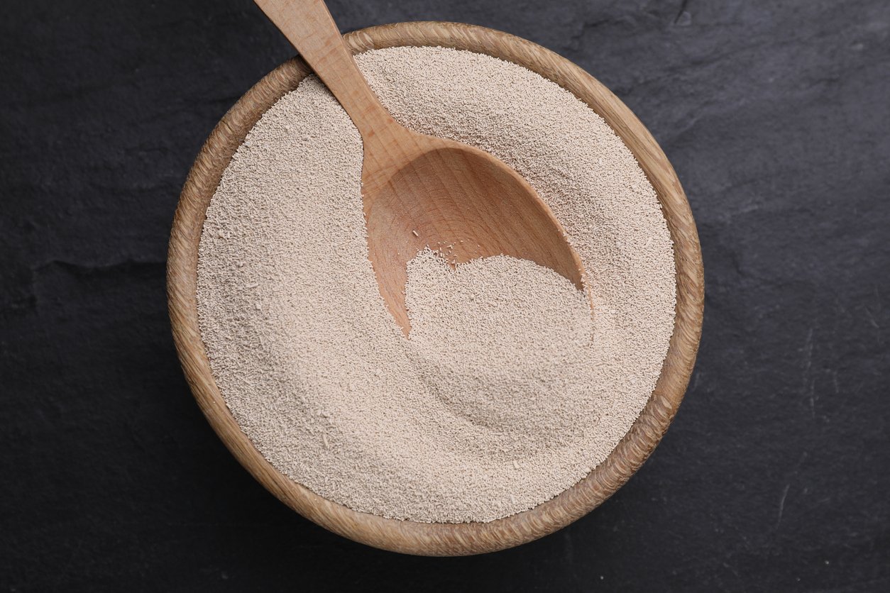 A wooden bowl filled with dry active yeast granules, with a wooden spoon resting inside, on a dark textured surface.