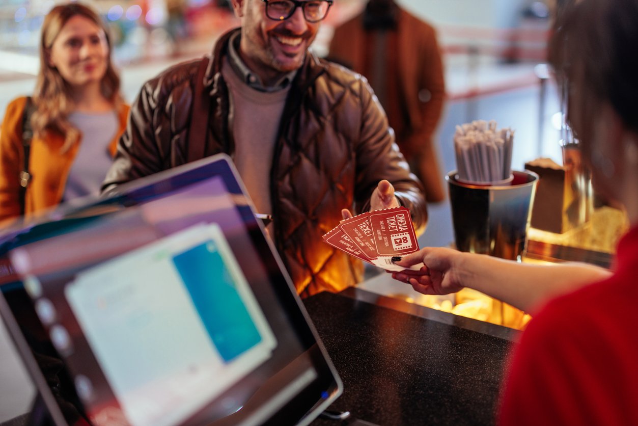 A smiling man hands movie tickets to an employee at a counter, while a woman waits behind him. A computer screen and containers with straws are visible in the foreground.