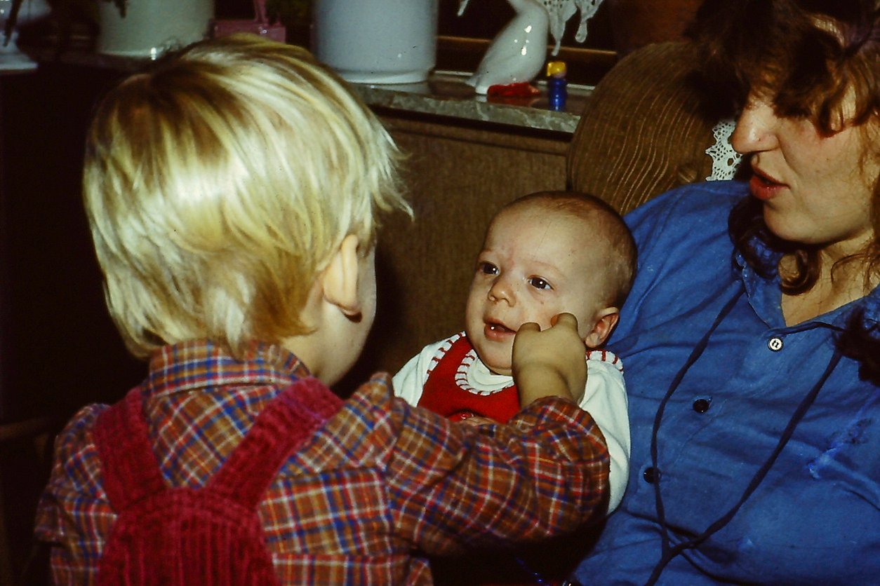 A young child in plaid and red overalls gently touches a baby’s cheek as the baby sits on a woman’s lap. The woman, wearing a blue shirt, looks at the children. Indoors, they share a tender moment before Thanksgiving dinner cost conversations begin.