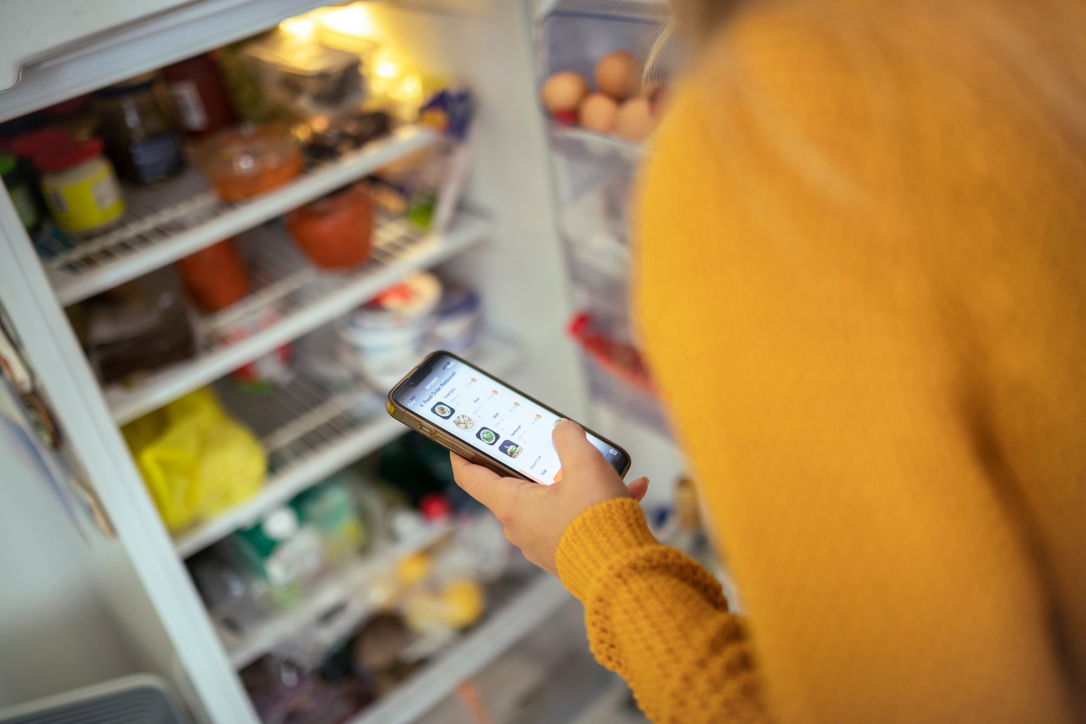 A person in a yellow sweater stands in front of an open refrigerator, holding a smartphone and looking at the screen, with various food items visible on the shelves inside the fridge.