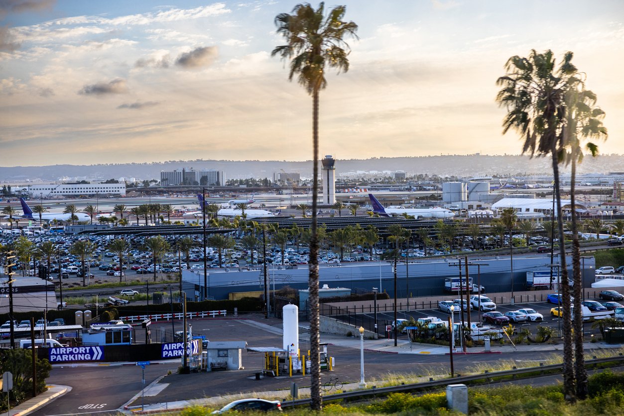 A busy airport with multiple airplanes, parking lots full of cars, and palm trees in the foreground under a partly cloudy sky at sunset.
