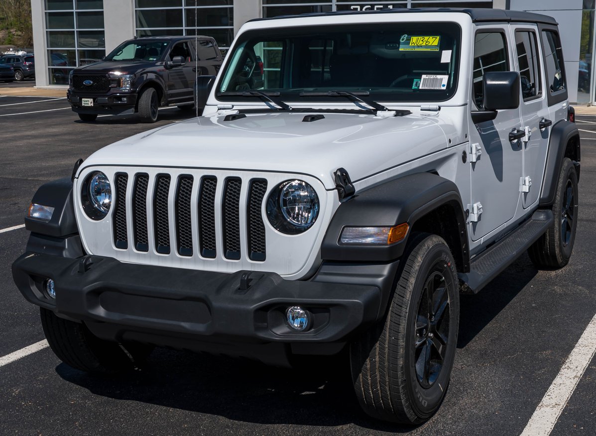 A white Jeep Wrangler SUV with black trim is parked in a lot outside a building, with another vehicle visible in the background.