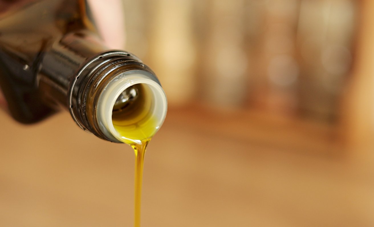 Close-up of olive oil being poured from a glass bottle, with a golden stream of oil flowing out. The background is softly blurred and warm-colored.
