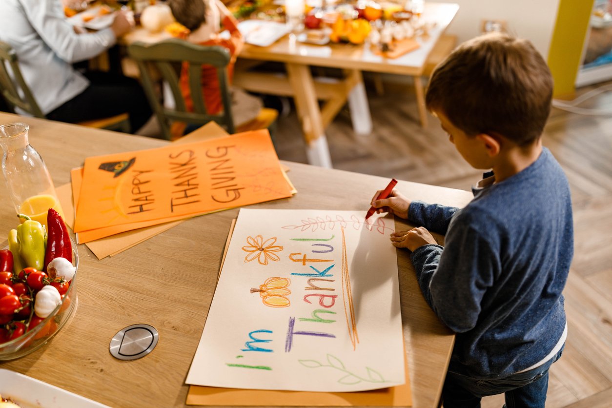 A young boy uses a marker to make a colorful "I'm Thankful" sign with pumpkins and flowers at a table, while others discuss Thanksgiving dinner cost in the background as they prepare for the holiday.