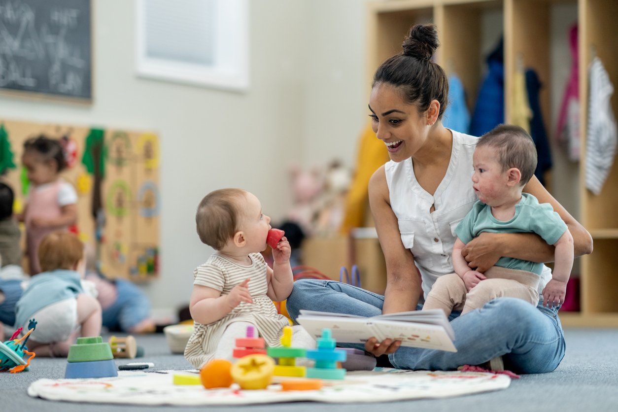 A woman sits cross-legged on the floor holding a baby and reading a book, while another baby sits nearby holding a red toy. Colorful toys are scattered on the mat in a lively daycare setting.