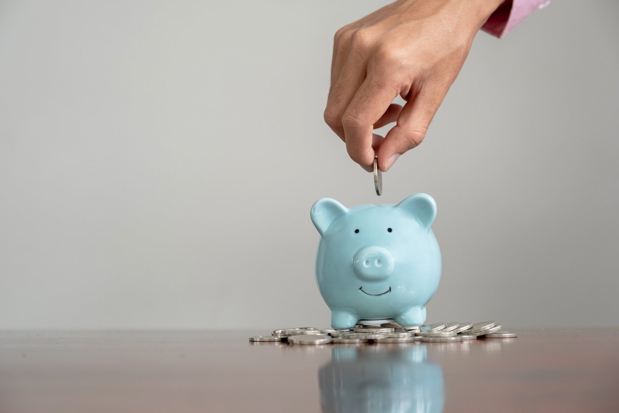 A hand places a coin into a blue piggy bank surrounded by scattered coins on a wooden surface, symbolizing saving money.