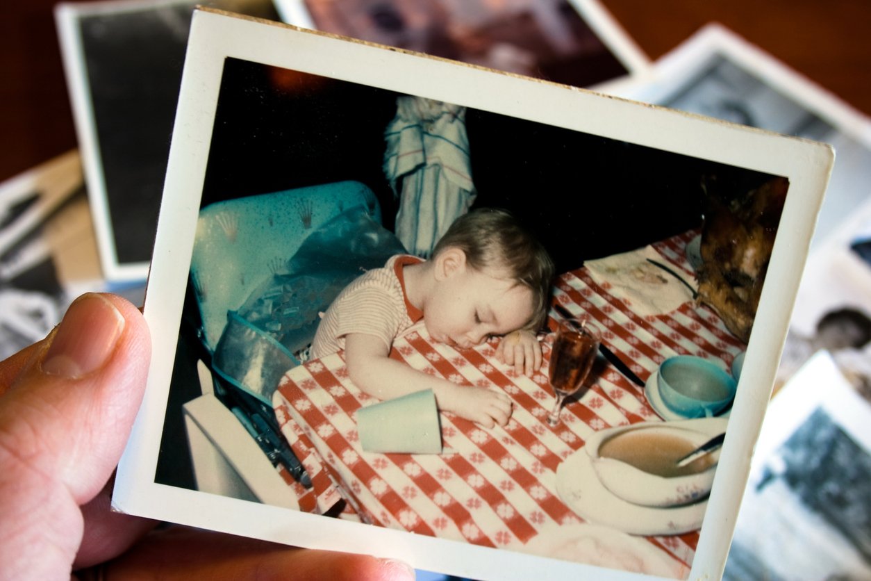 A hand holds a photo of a small child asleep at a table with a red and white checkered tablecloth, surrounded by dishes, a cup, and other photos—perhaps from gatherings over the years—reflecting on Thanksgiving Dinner Cost History.