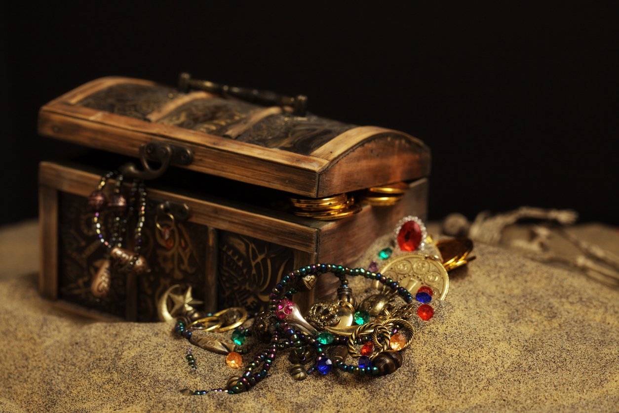 An open wooden treasure chest filled with gold coins, necklaces, and colorful jewels sits on sand, with some jewelry spilling out onto the ground.