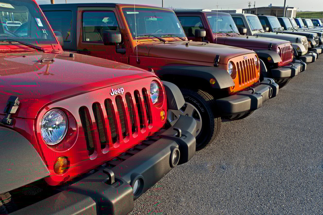 A row of brightly colored Jeep Wranglers parked side by side in a lot on a sunny day, with the focus on the front grills and headlights.