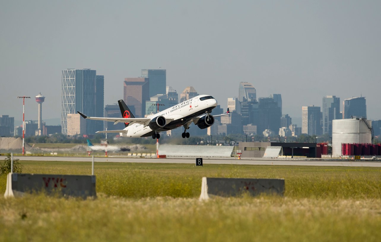 An Air Canada airplane takes off from a runway with the city skyline and skyscrapers visible in the background under a clear sky.