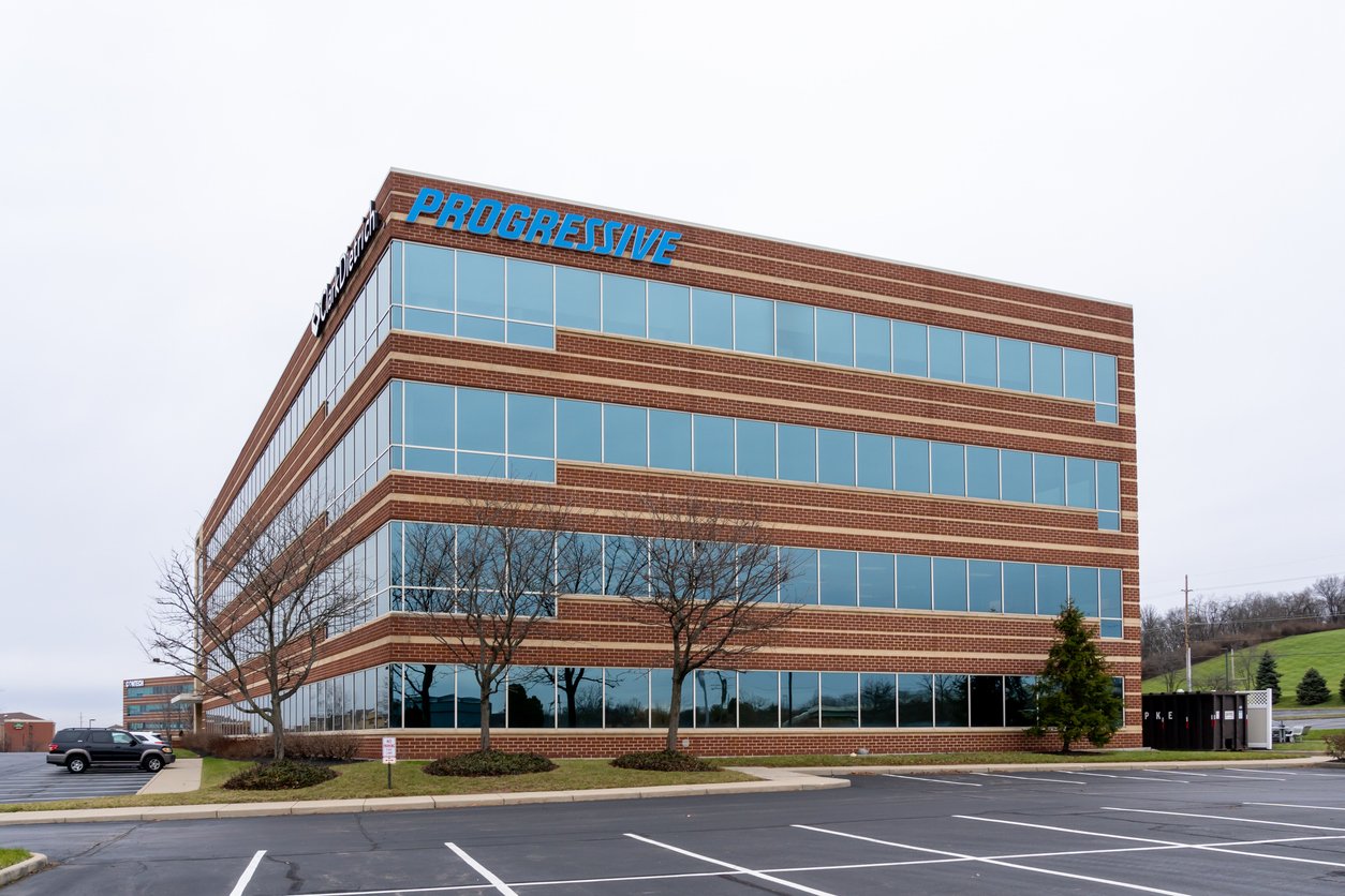 A modern brick office building with large blue-tinted windows and the "Progressive" sign on top, surrounded by a mostly empty parking lot and a few leafless trees, under an overcast sky.