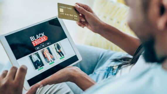 A person holding a credit card shops online for Black Friday deals on a tablet, browsing clothing items with sale prices displayed on the screen.