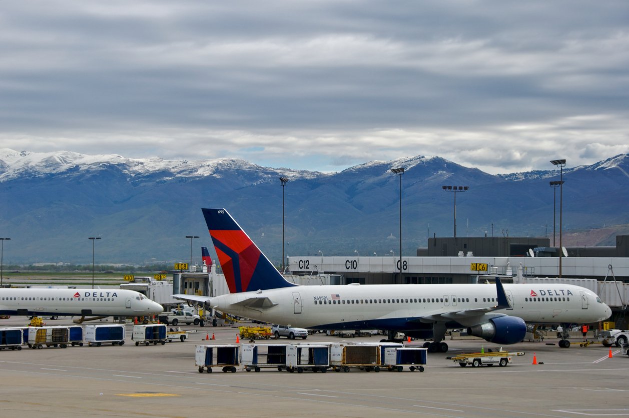 Delta Airlines planes parked at airport gates with baggage carts nearby, and snow-capped mountains visible in the background under a cloudy sky.