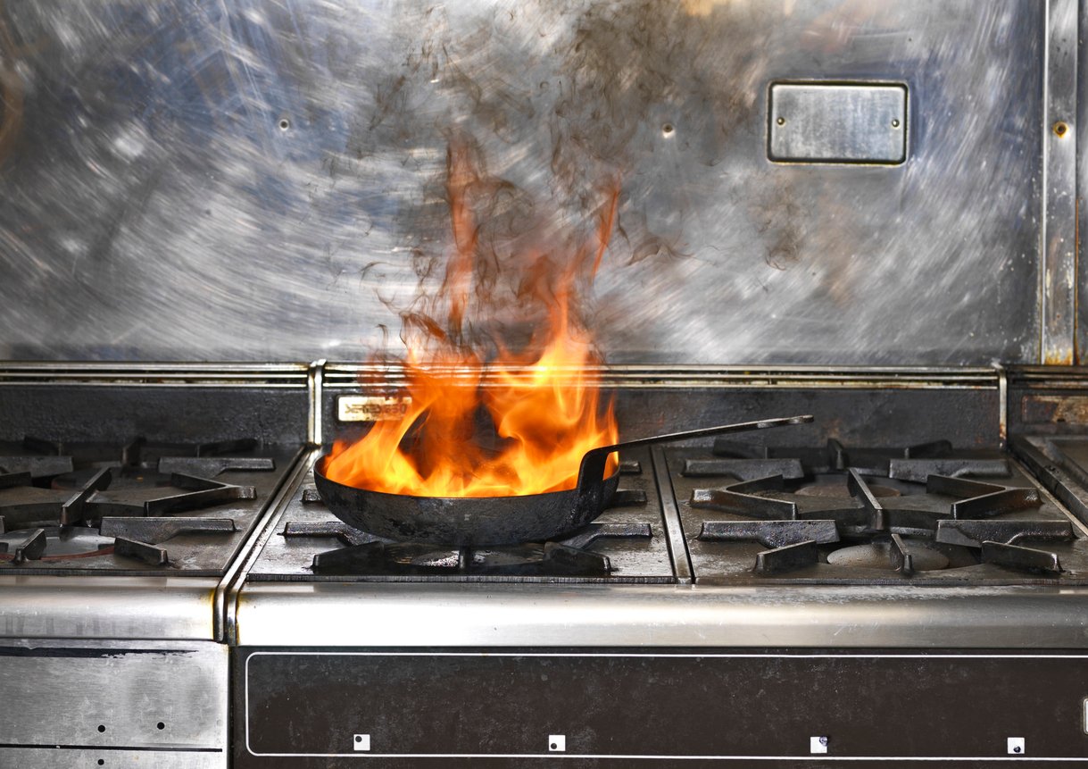A metal frying pan on a stove with large orange flames rising from it, indicating a kitchen fire. The stove and surrounding area are made of stainless steel.
