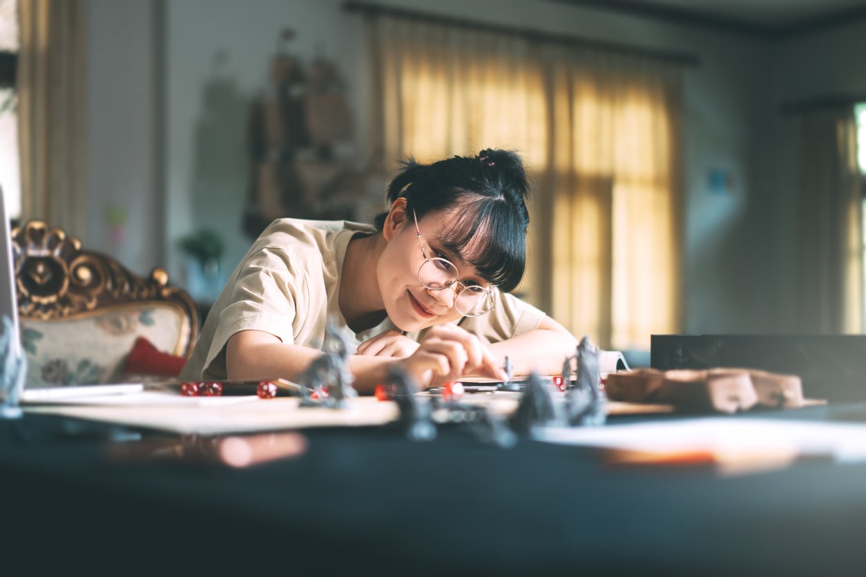 A young person with glasses smiles while playing a tabletop game at a sunny indoor table, surrounded by game pieces, dice, and papers.