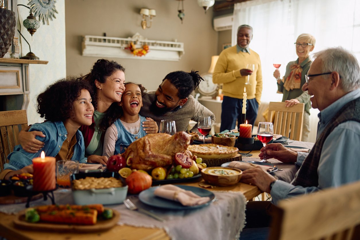 A multigenerational family gathers around a festive dinner table, laughing and hugging. As they enjoy the feast, conversations spark about thanksgiving dinner cost, with turkey, candles, and side dishes filling the table. Two people stand in the background, smiling with drinks.