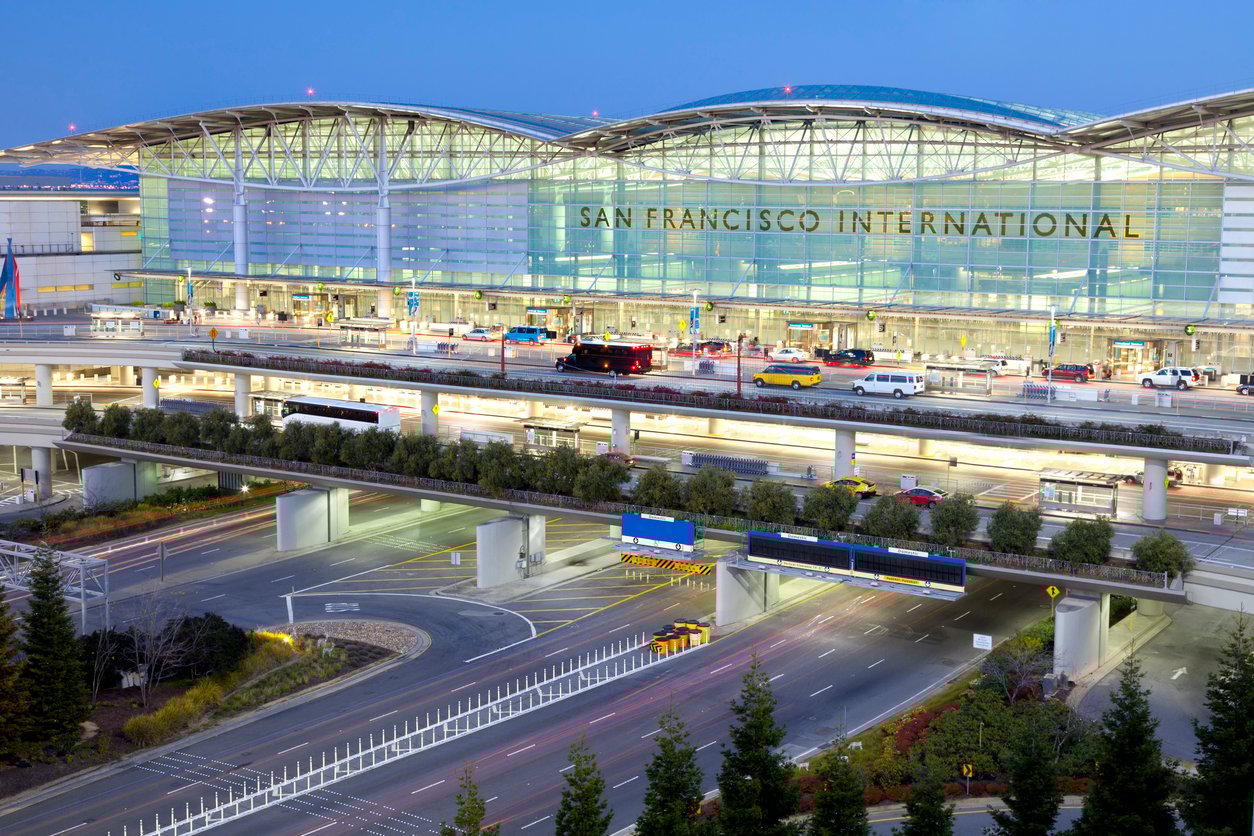 San Francisco International Airport exterior at dusk, showing a large glass terminal building, multiple levels of roads, cars, buses, and trees in the foreground.