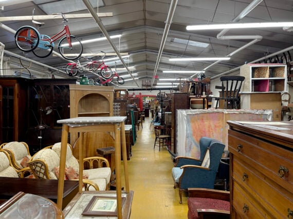 A cluttered thrift store or antique shop interior with assorted vintage furniture, bicycles hanging from the ceiling, stacked chairs, and shelves filled with various household items along a bright yellow floor.