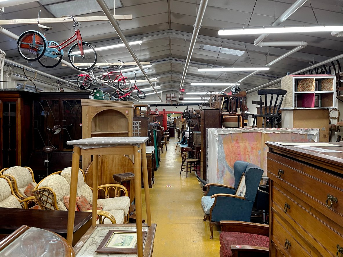 A cluttered thrift store or antique shop interior with assorted vintage furniture, bicycles hanging from the ceiling, stacked chairs, and shelves filled with various household items along a bright yellow floor.