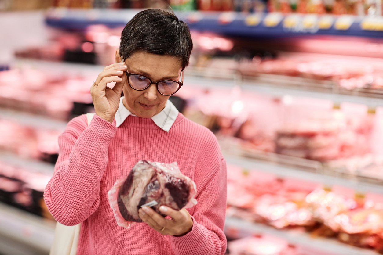 A person wearing glasses and a pink sweater examines a package of raw meat while shopping in the meat section of a grocery store. Shelves with various meats are visible in the background.