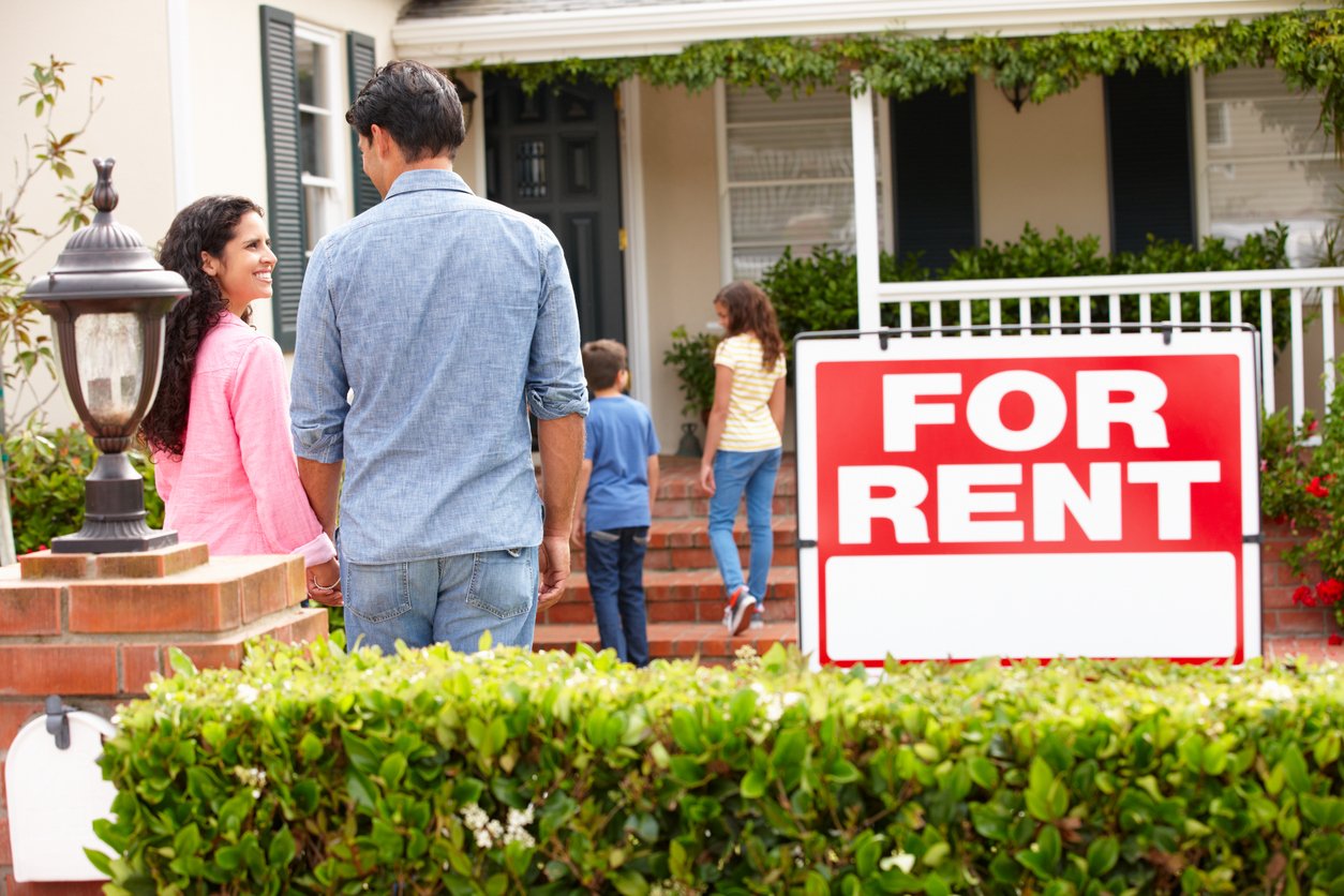 A family with two children walks toward a house with a porch, passing a large "For Rent" sign in the yard. The parents smile at each other, and the kids head up the steps.