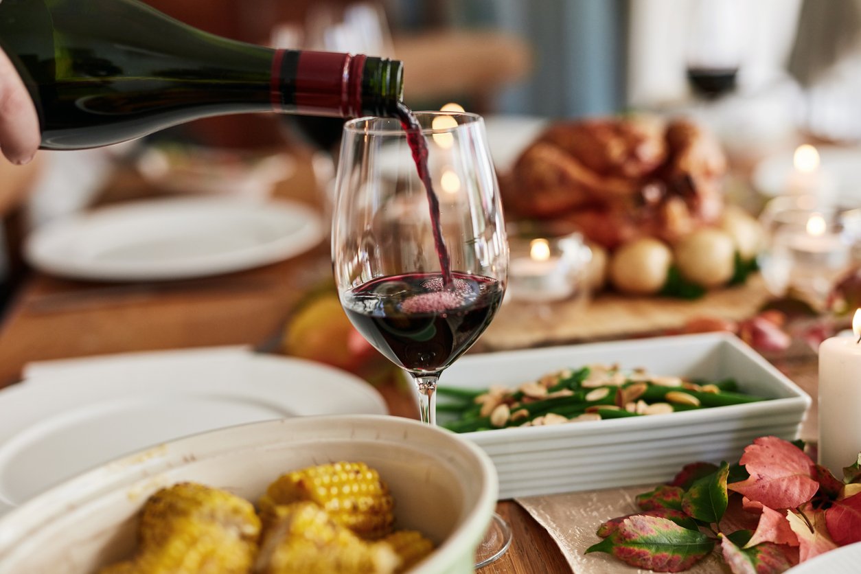 A hand pours red wine into a glass on a festive table set with corn, green beans, and a roast, surrounded by candles and autumn leaves.