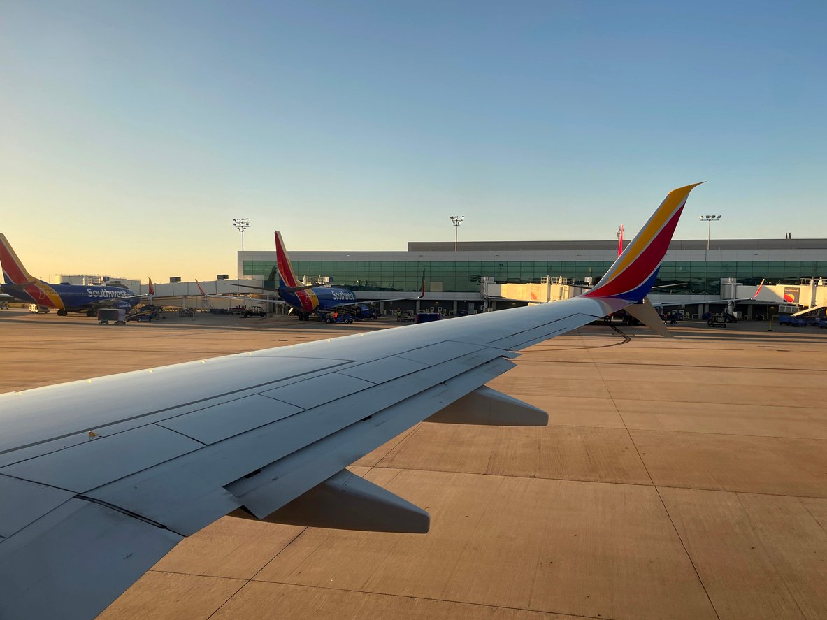 View from an airplane window showing the wing, airport tarmac, and several Southwest Airlines planes parked near the terminal under a clear sky during daylight.