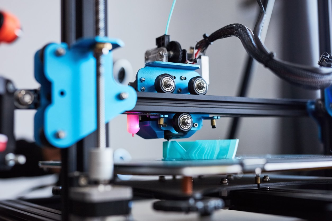 Close-up of a 3D printer in operation, with the extruder head depositing blue filament onto the print bed, creating a partially completed object.