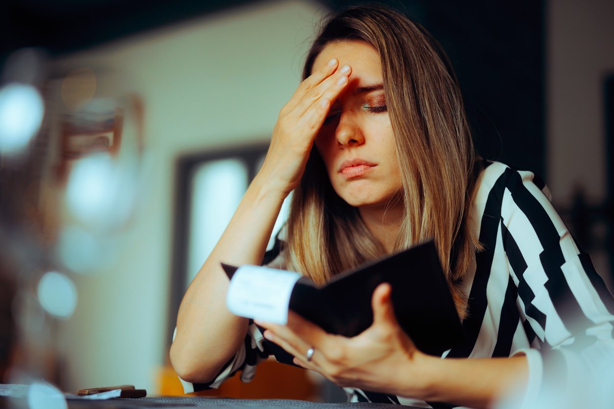 A woman with long hair sits at a table, holding her forehead with one hand and a wallet in the other, looking stressed or worried.