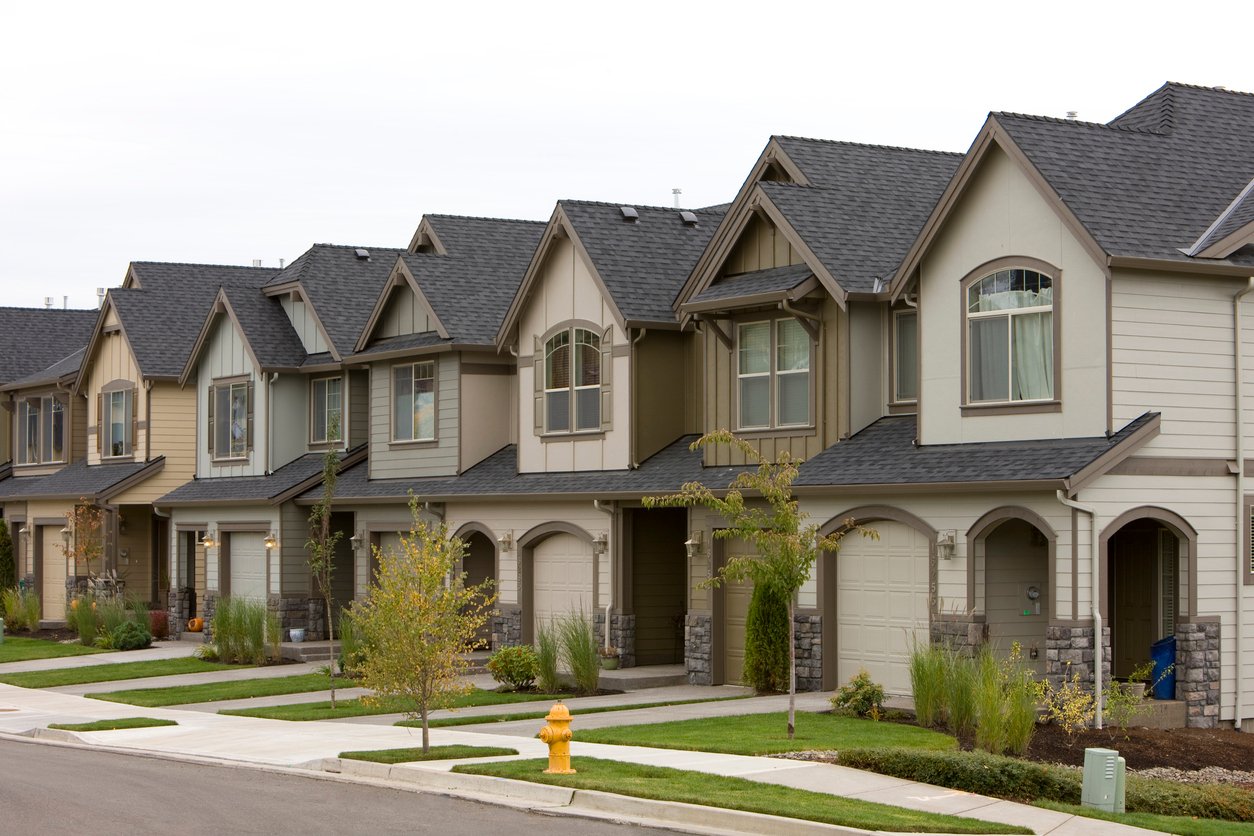 A row of modern, two-story suburban houses with garages, front lawns, and young trees lines a quiet residential street. A yellow fire hydrant stands on the sidewalk. The sky is overcast.