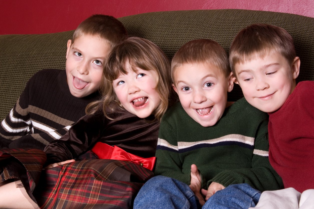 Four young children sit closely together on a couch, smiling and making silly faces at the camera. Dressed in sweaters and plaid or velvet clothes, they bring warmth reminiscent of Thanksgiving dinner cost celebrations, set against a red wall in the background.