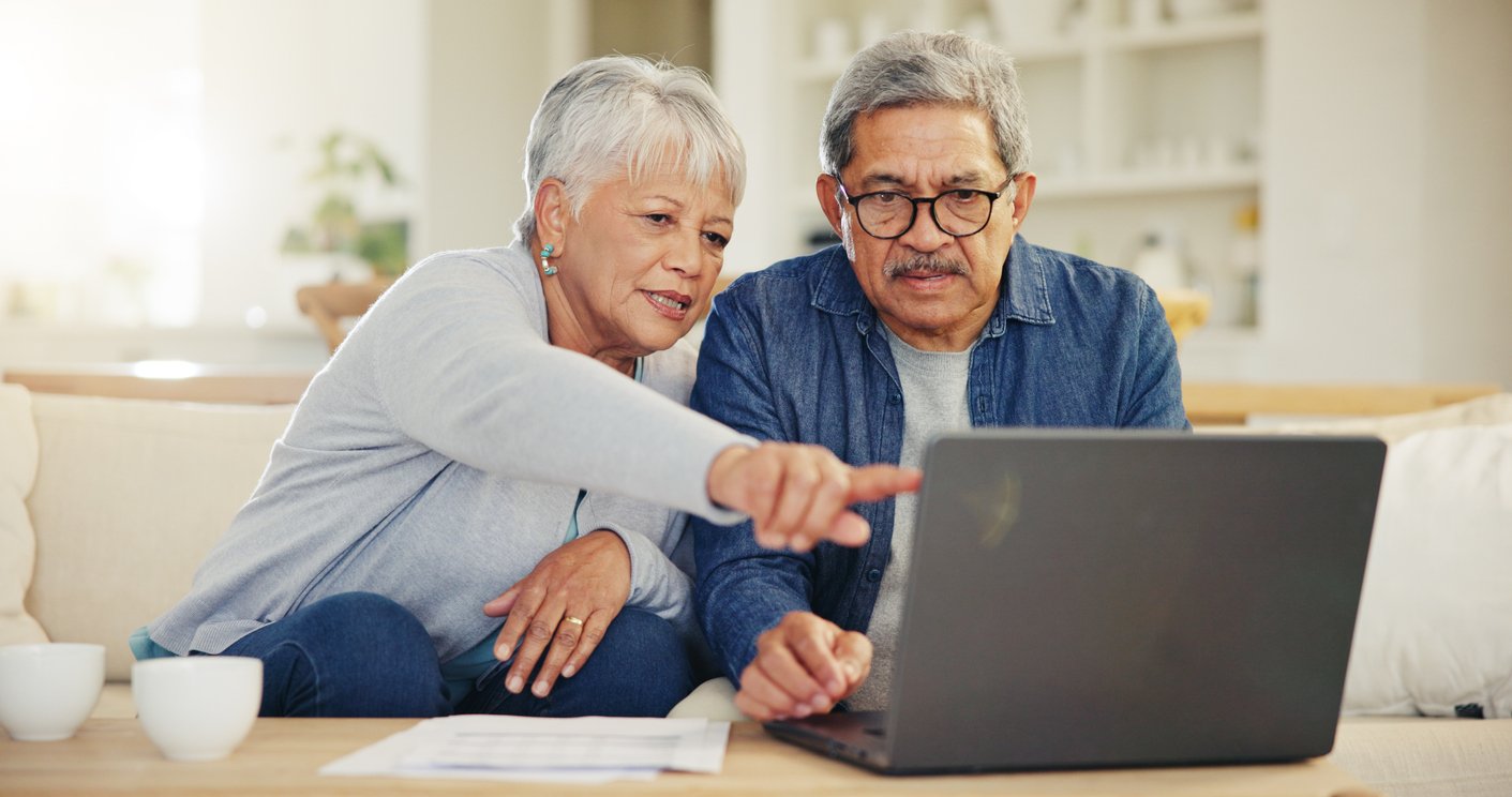 An elderly woman and man sit on a couch, looking intently at a laptop. The woman points at the screen while the man types, both appearing focused and engaged. Papers and coffee cups are on the table in front of them.