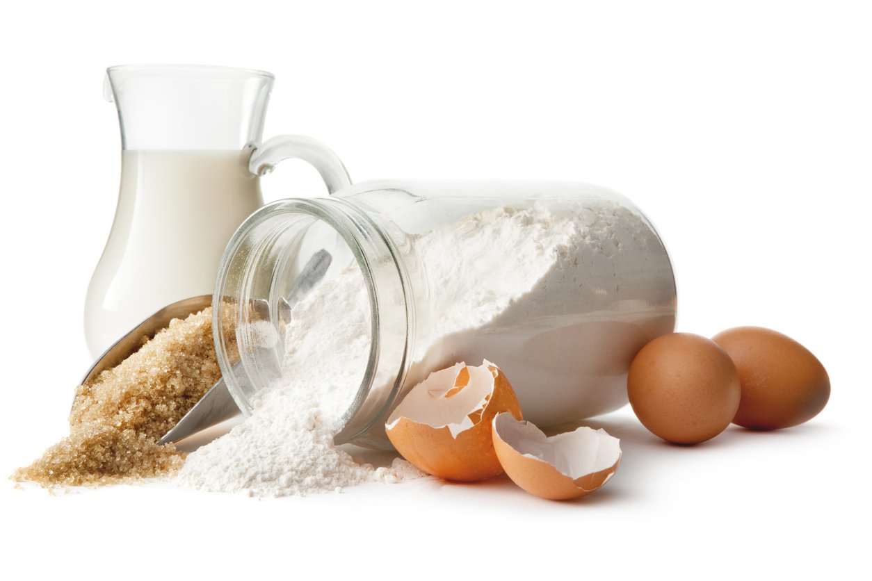 A glass jar of spilled flour, brown sugar with a scoop, a small glass pitcher of milk, three whole eggs, and one cracked eggshell, all arranged on a white background.