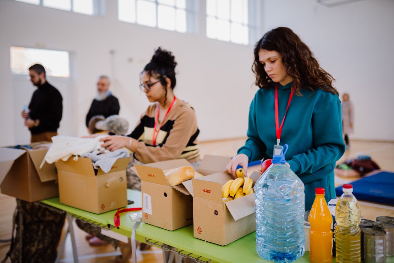 Two women pack food and clothing items into cardboard boxes on a table in a bright room, preparing supplies. Bottles of water, juice, and various groceries are visible. People stand and work in the background.