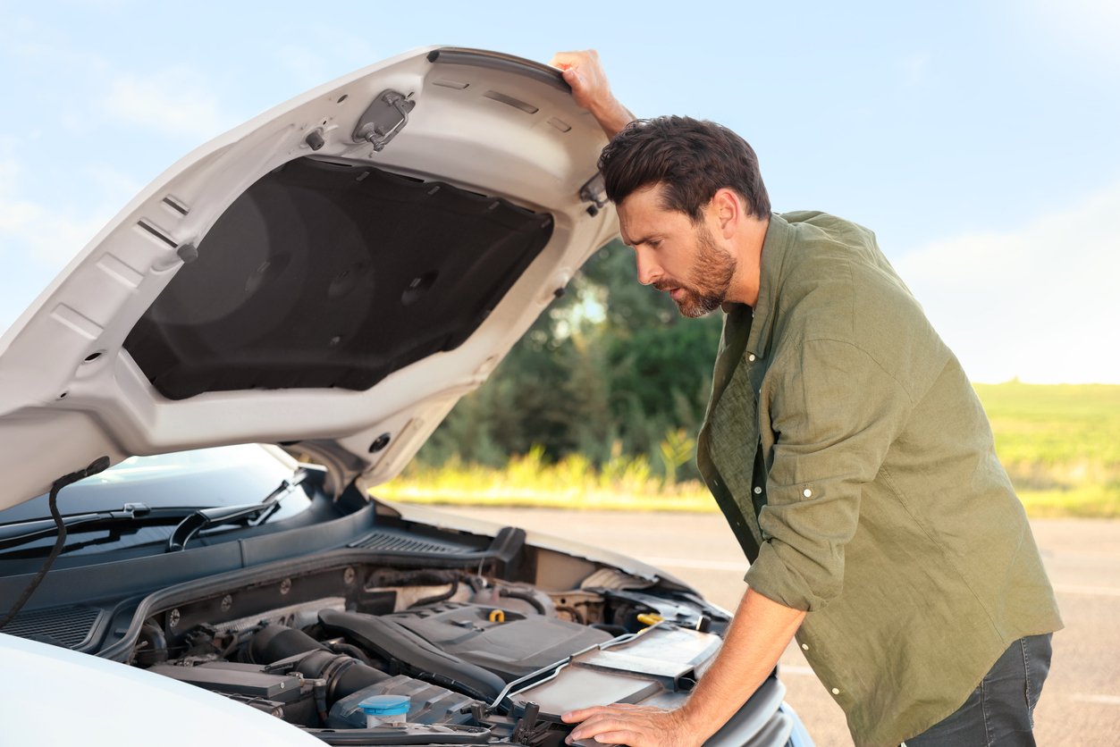 A man standing outdoors looks concerned while examining the engine of a white car with the hood open. Trees and a blue sky are visible in the background.