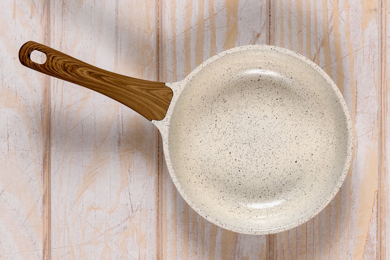 A ceramic frying pan with a speckled white interior and a wooden handle sits on a light wooden surface, viewed from above.