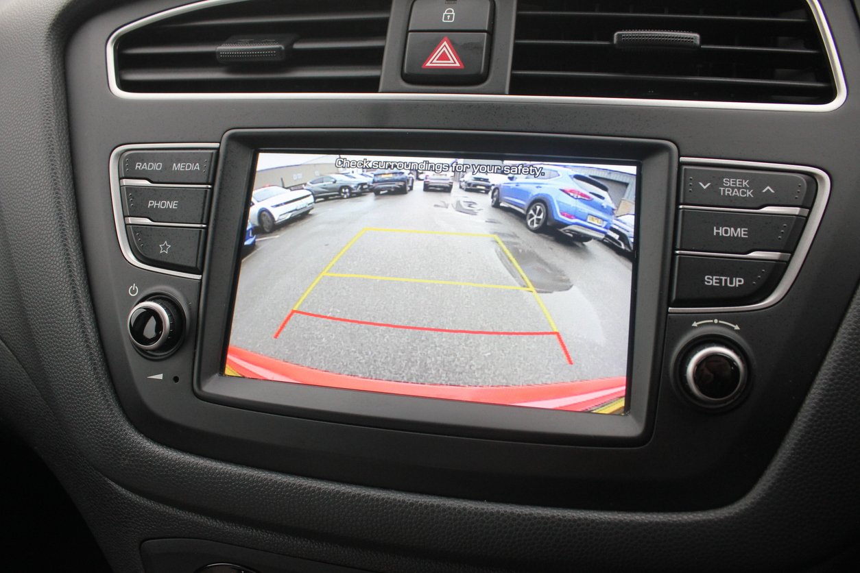 A car's dashboard display shows the view from the rearview camera, with colored guide lines and parked cars visible behind, helping the driver reverse safely.