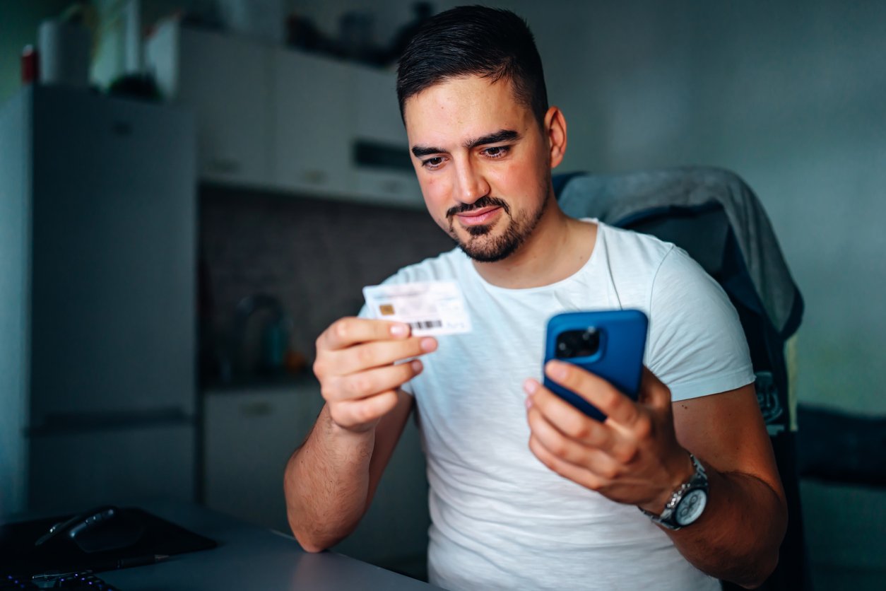 A man sits at a desk in a kitchen, holding a smartphone in one hand and an ID card in the other, appearing focused as he looks at the ID card. He wears a white t-shirt and a wristwatch.