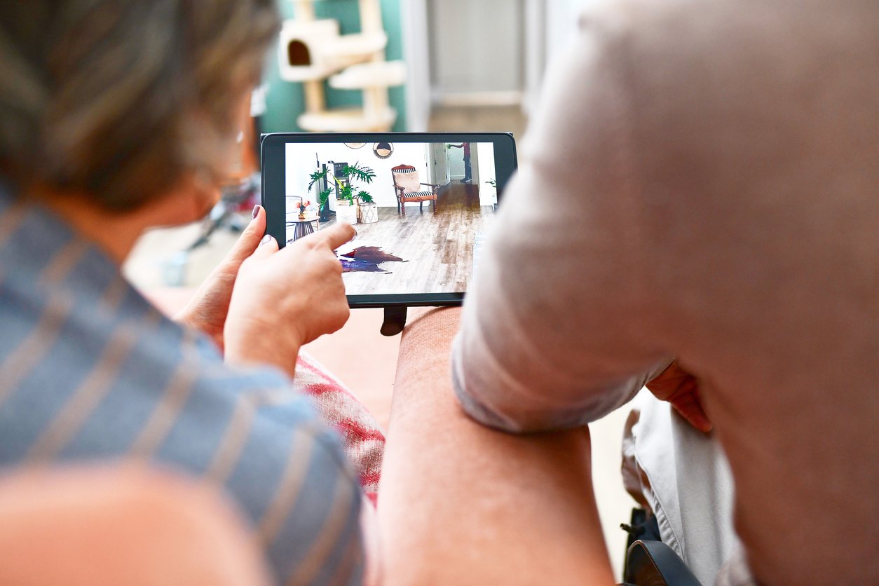 Two people sit closely together, viewing a tablet showing a home interior with a dog lying on a wooden floor. One person is pointing at the screen, and their faces are not visible in the image.