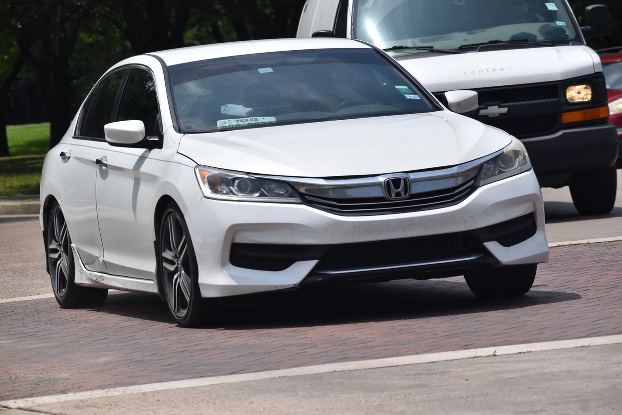 A white Honda sedan driving on a street, with a white Chevrolet van visible behind it. The cars appear to be in motion, and trees can be seen in the background.