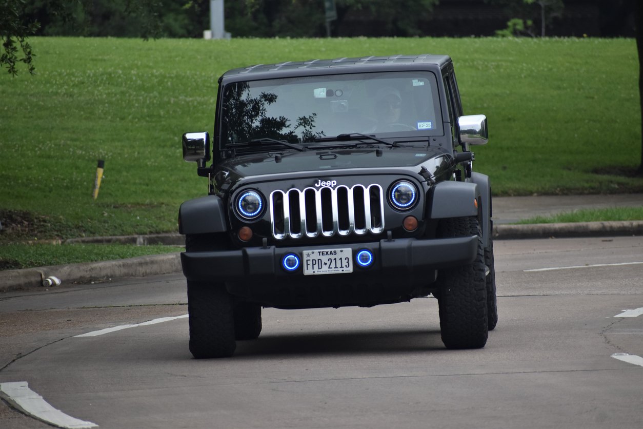A black Jeep Wrangler with Texas license plates drives on a curved road bordered by grass and trees. Two people are visible inside the vehicle. The headlights have blue rings around them.