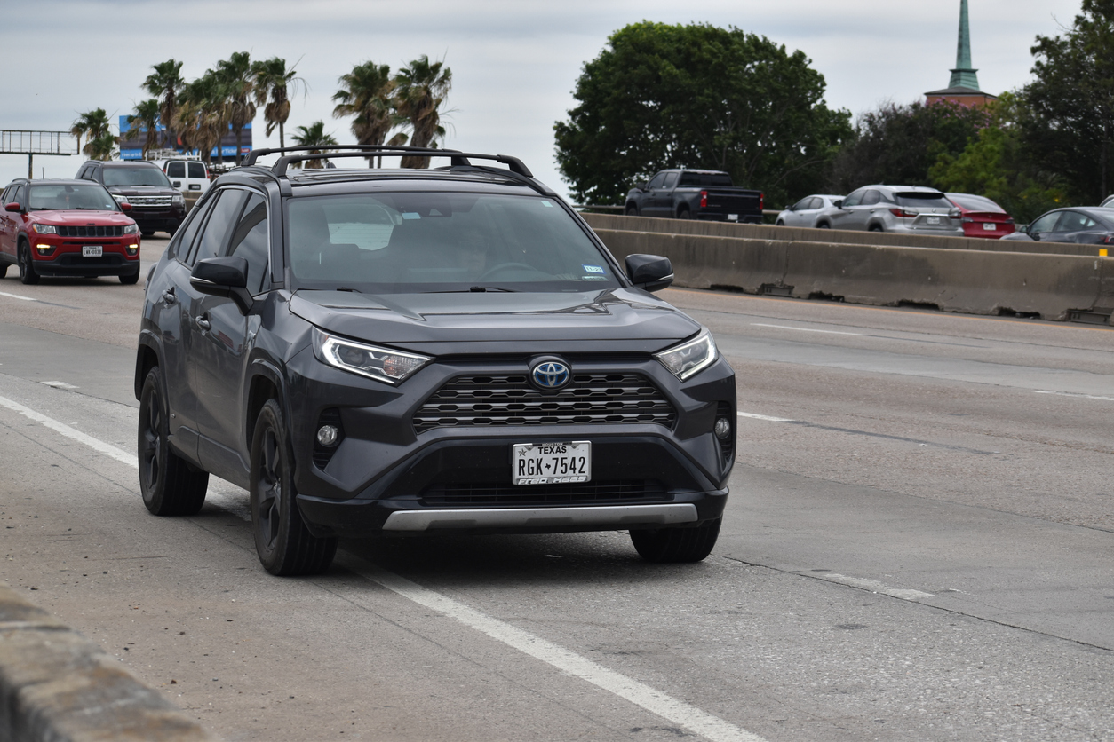 A gray Toyota RAV4 with Texas license plates is parked on the shoulder of a busy highway, with palm trees, other vehicles, and a church steeple visible in the background.