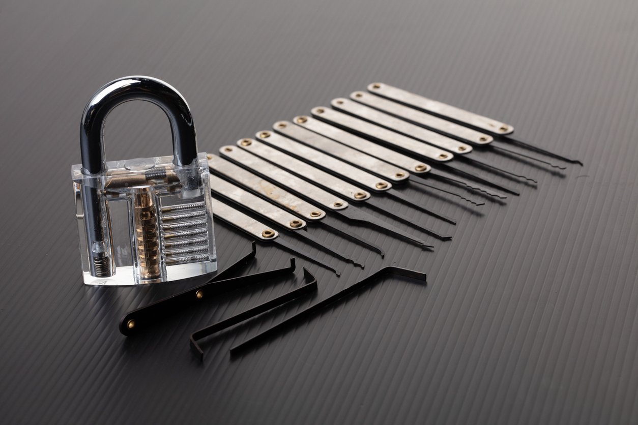 A transparent padlock and multiple lock picking tools are arranged on a dark, textured surface, showing the inner mechanisms of the lock and various picks for manipulating it.
