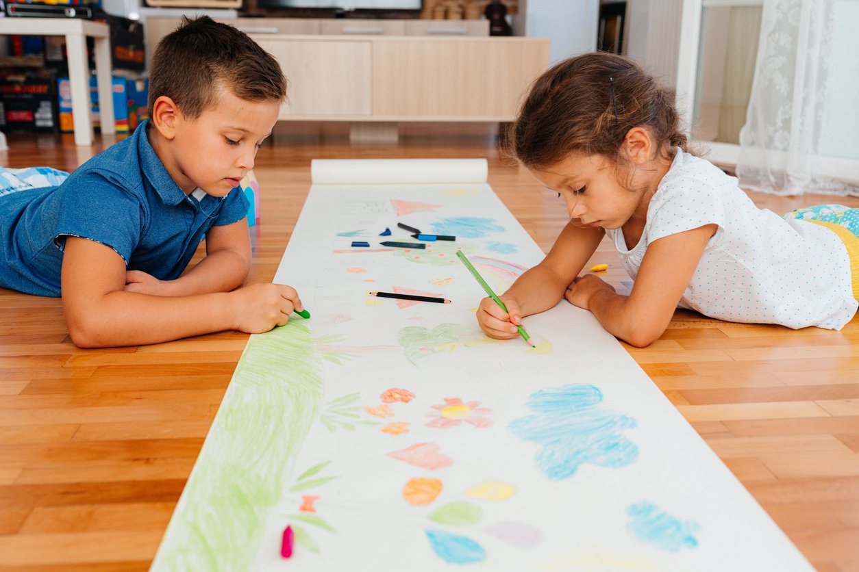 Two young children are lying on a wooden floor, drawing with colored pencils and crayons on a long sheet of white paper filled with colorful flowers and shapes. They are focused and engaged in their artwork.