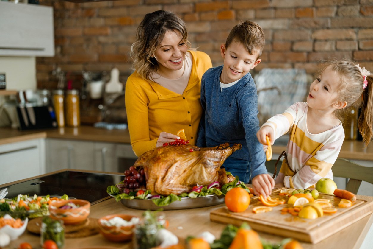 A woman and two children prepare a festive meal in a kitchen, smiling as they decorate a large roasted turkey, reflecting on Thanksgiving dinner cost history while arranging fruits and vegetables on the counter.