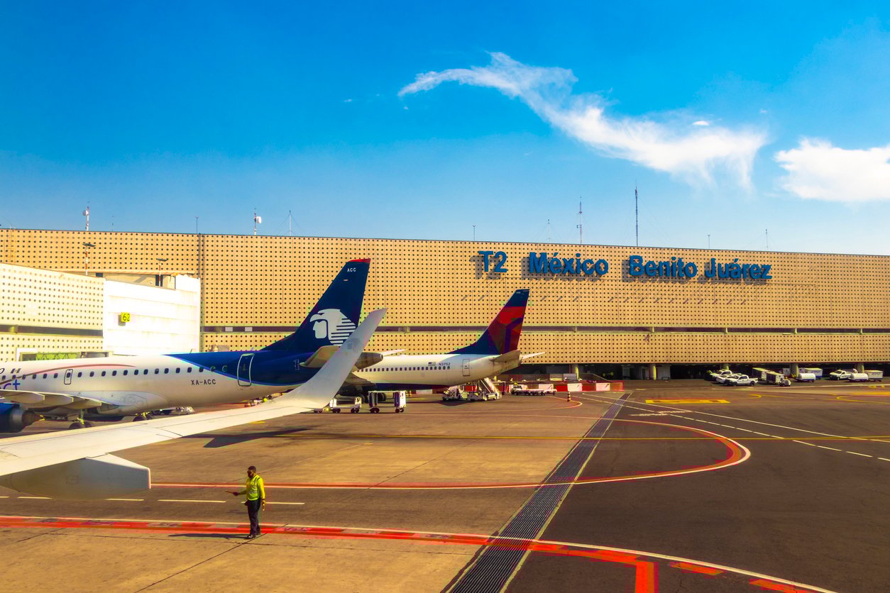 A view of Terminal 2 at Mexico City’s Benito Juárez International Airport, showing parked airplanes, a person in a high-visibility vest on the tarmac, and clear blue sky above.