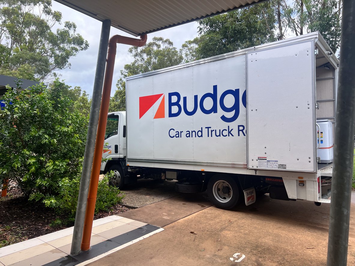 A white Budget rental truck is parked near a building surrounded by greenery. The truck has the Budget logo and "Car and Truck Rental" written on its side. Part of a roof and drainage pipe are visible in the foreground.