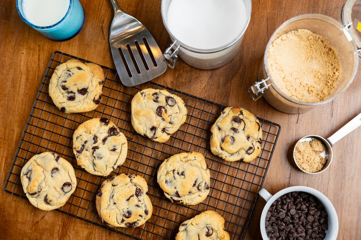 Chocolate chip cookies cooling on a wire rack, surrounded by ingredients including chocolate chips, brown sugar, flour, a cup of milk, and a metal spatula on a wooden surface.