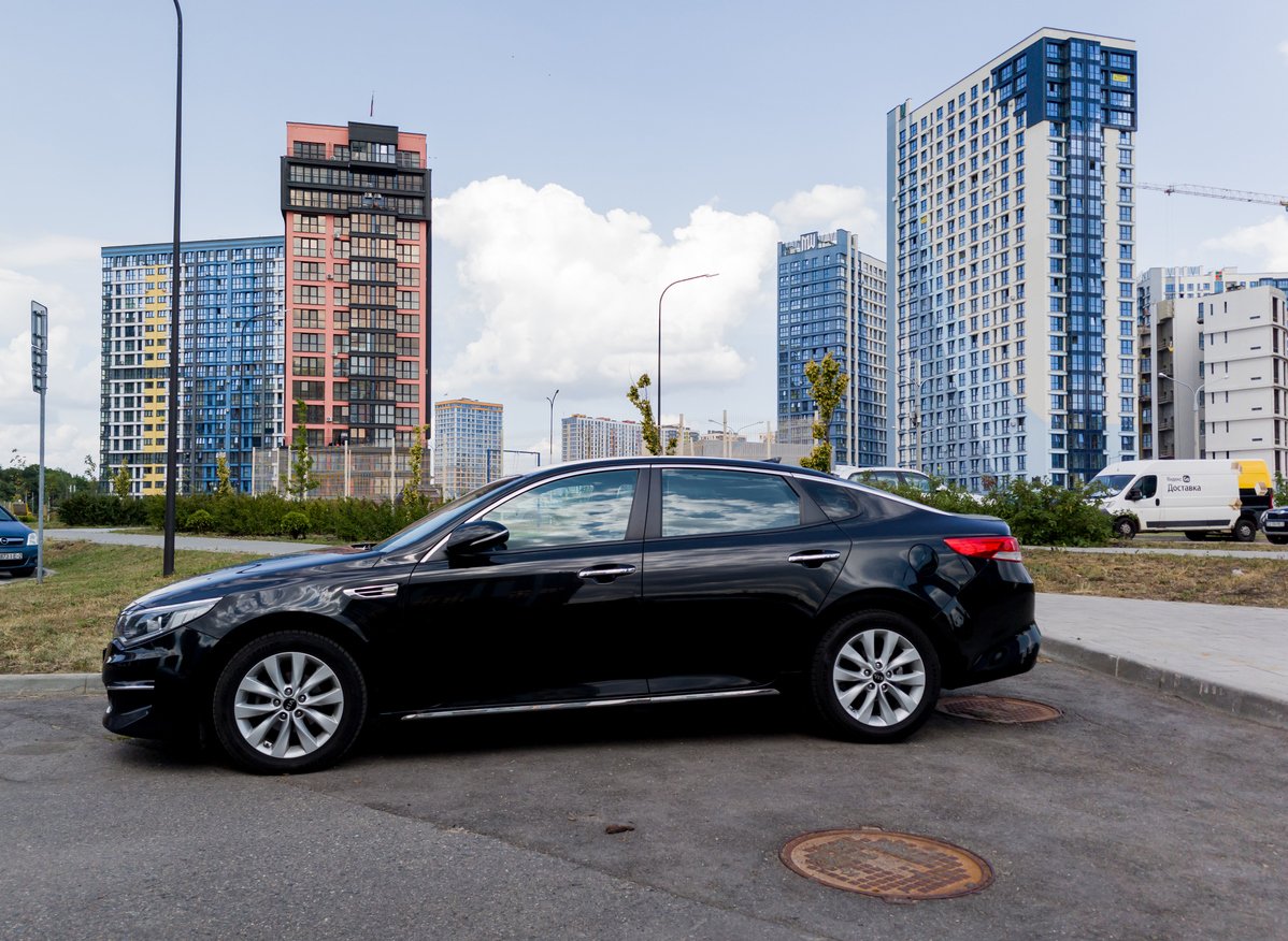 A black sedan is parked on a paved area near modern high-rise apartment buildings under a partly cloudy sky. Some greenery and parked vehicles are visible in the background.