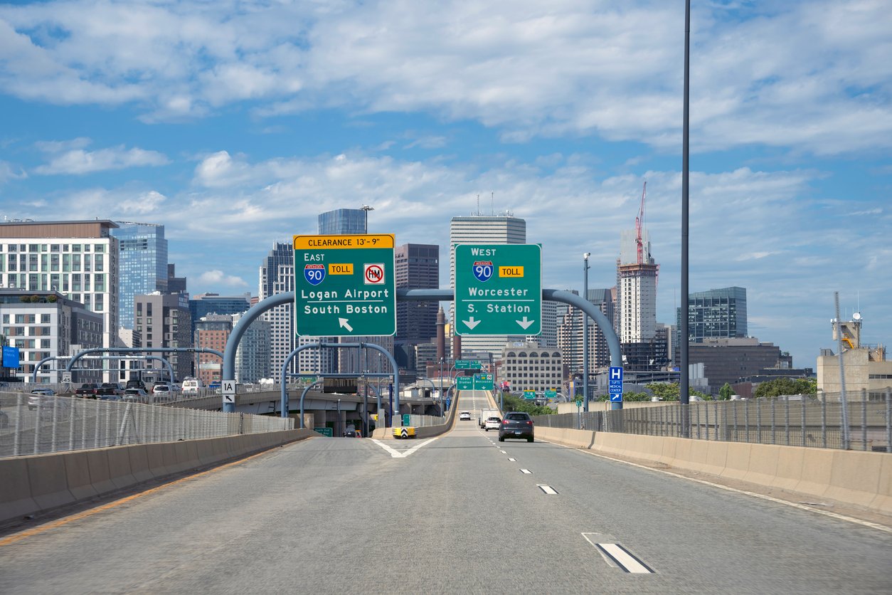 A view from a highway approaching downtown Boston, showing signs for I-90 East to Logan Airport and South Boston, and I-90 West to Worcester, with cars and city buildings visible ahead under a partly cloudy sky.